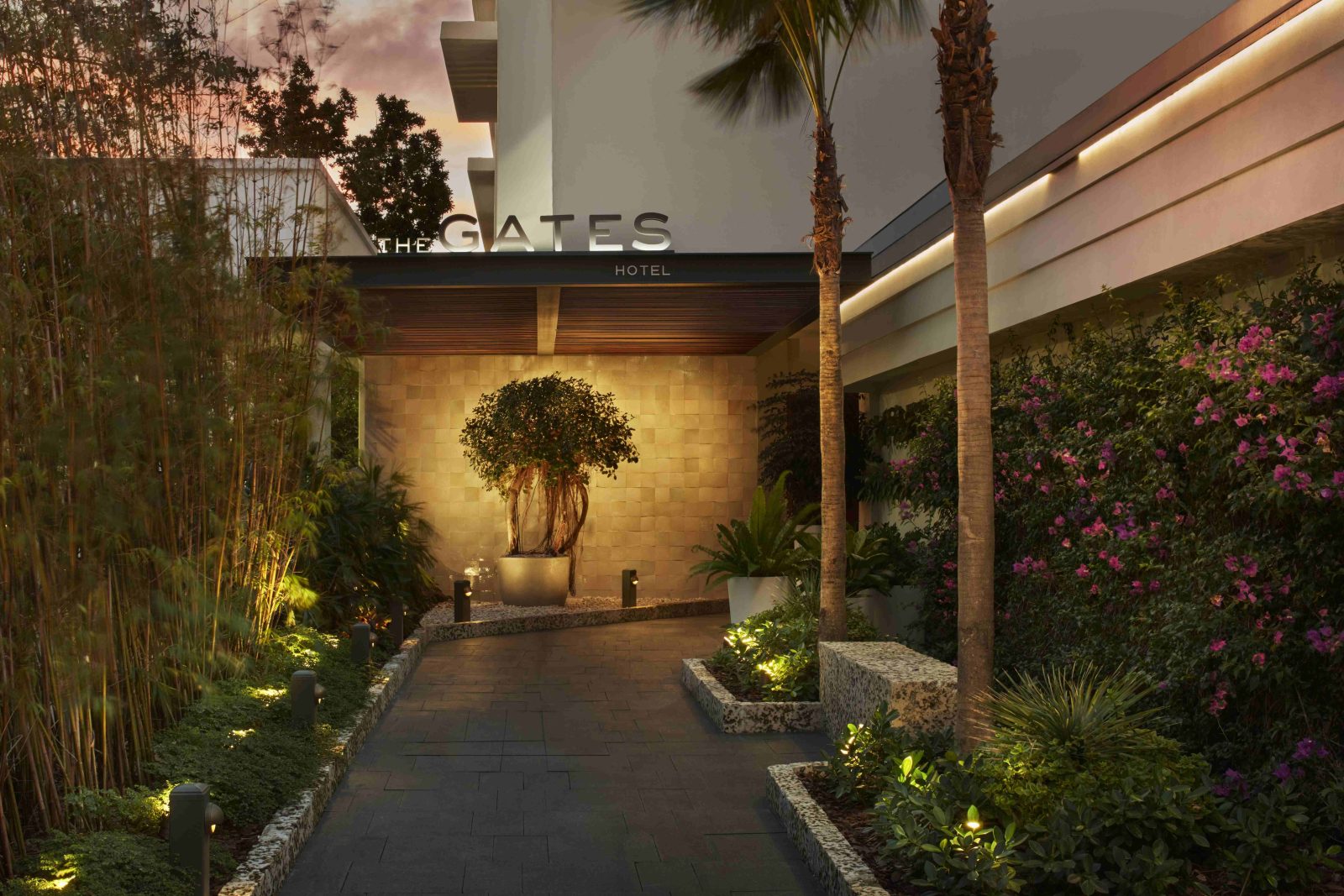 Illuminated entrance to The Gates Hotel South Beach with lush greenery and evening ambiance. Caption: The Gates Hotel welcomes guests through a serene, tropical-inspired entrance just steps from the beach.