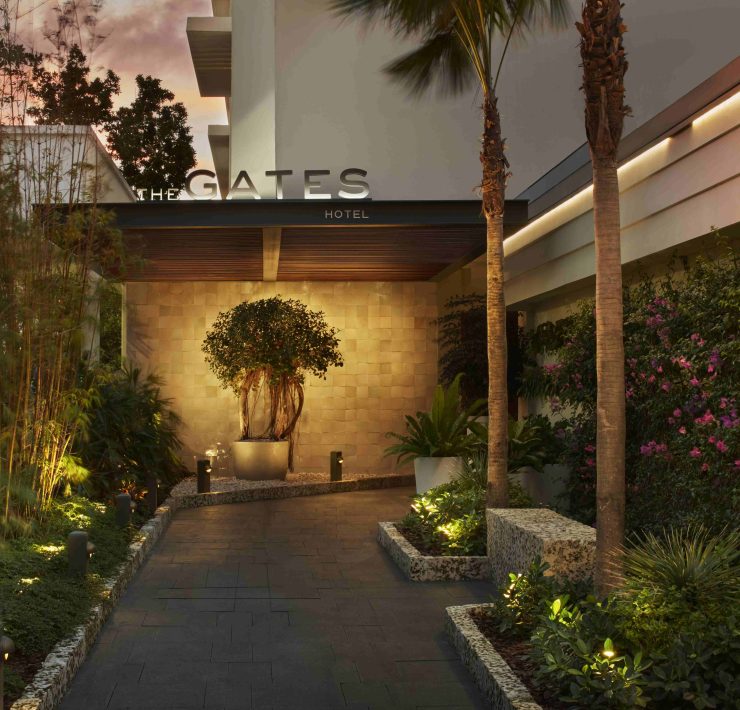 Illuminated entrance to The Gates Hotel South Beach with lush greenery and evening ambiance. Caption: The Gates Hotel welcomes guests through a serene, tropical-inspired entrance just steps from the beach.
