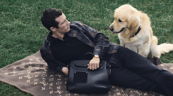 Man lying on grass with golden retriever and Louis Vuitton LV Touch black leather bag.