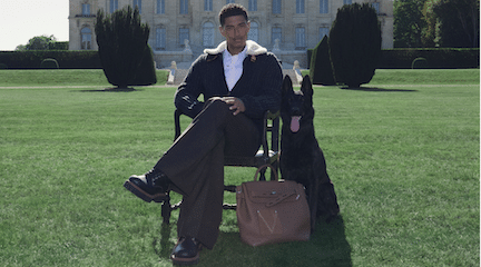 Model sitting on a chair with black dog and Louis Vuitton LV Touch Steamer bag on green lawn.