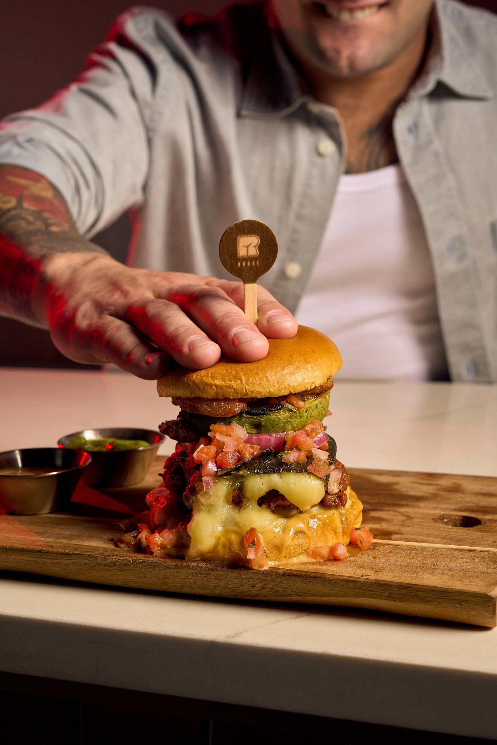 Close-up of a gourmet burger from R House Wynwood topped with crispy fritas, melted cheese, and pickles on a wooden board.