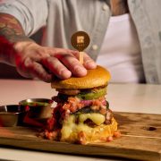 Close-up of a gourmet burger from R House Wynwood topped with crispy fritas, melted cheese, and pickles on a wooden board.