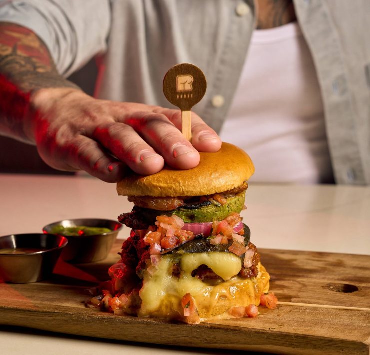 Close-up of a gourmet burger from R House Wynwood topped with crispy fritas, melted cheese, and pickles on a wooden board.