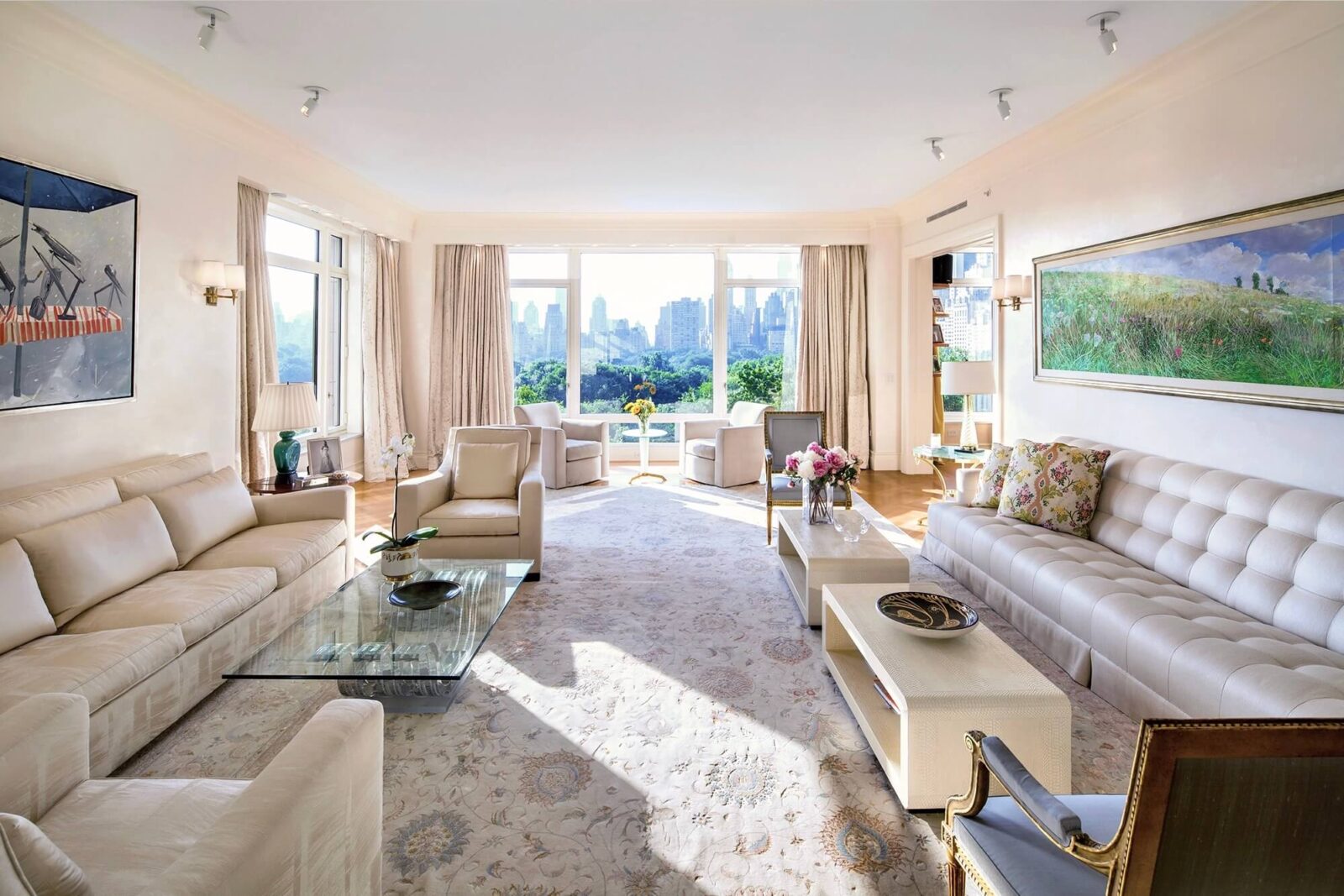 Sunlit living room with cream sofas, floral rug, glass coffee table, and expansive city skyline views