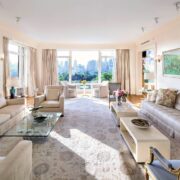 Sunlit living room with cream sofas, floral rug, glass coffee table, and expansive city skyline views