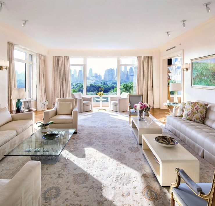Sunlit living room with cream sofas, floral rug, glass coffee table, and expansive city skyline views