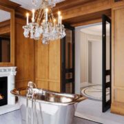 Luxury bathroom with warm wood paneling, crystal chandelier, freestanding metal tub, and marble fireplace