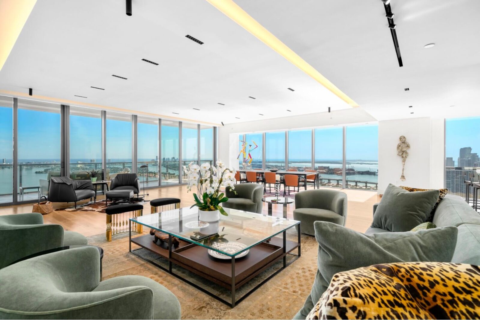 Open-concept penthouse living room with panoramic water views, soft green seating, and expansive floor-to-ceiling glass
