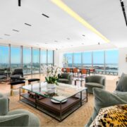 Open-concept penthouse living room with panoramic water views, soft green seating, and expansive floor-to-ceiling glass