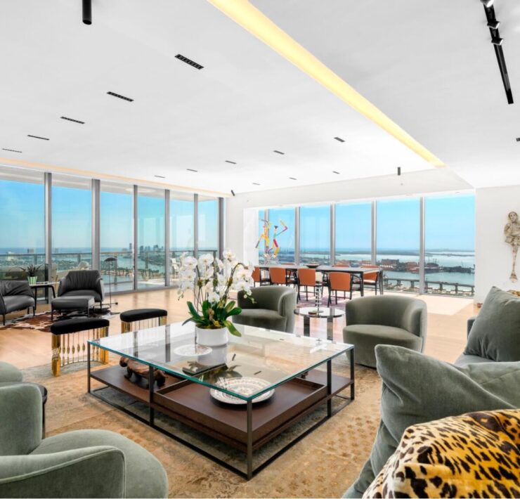 Open-concept penthouse living room with panoramic water views, soft green seating, and expansive floor-to-ceiling glass