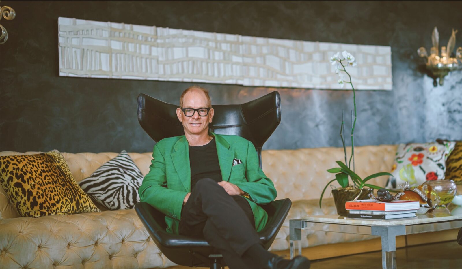Portrait of designer Kevin Gray seated in a black lounge chair, wearing a green blazer in a bold, art-filled interior with patterned pillows and dark walls