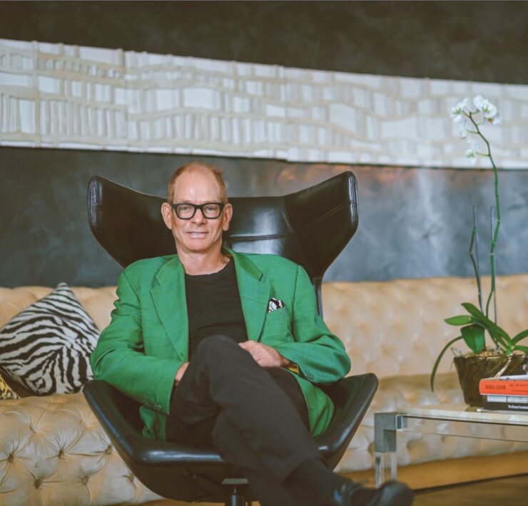 Portrait of designer Kevin Gray seated in a black lounge chair, wearing a green blazer in a bold, art-filled interior with patterned pillows and dark walls