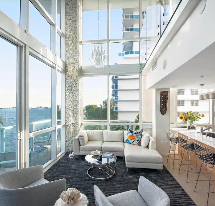 Double-height waterfront living space with white sectional, glass walls, modern kitchen island, and bay views