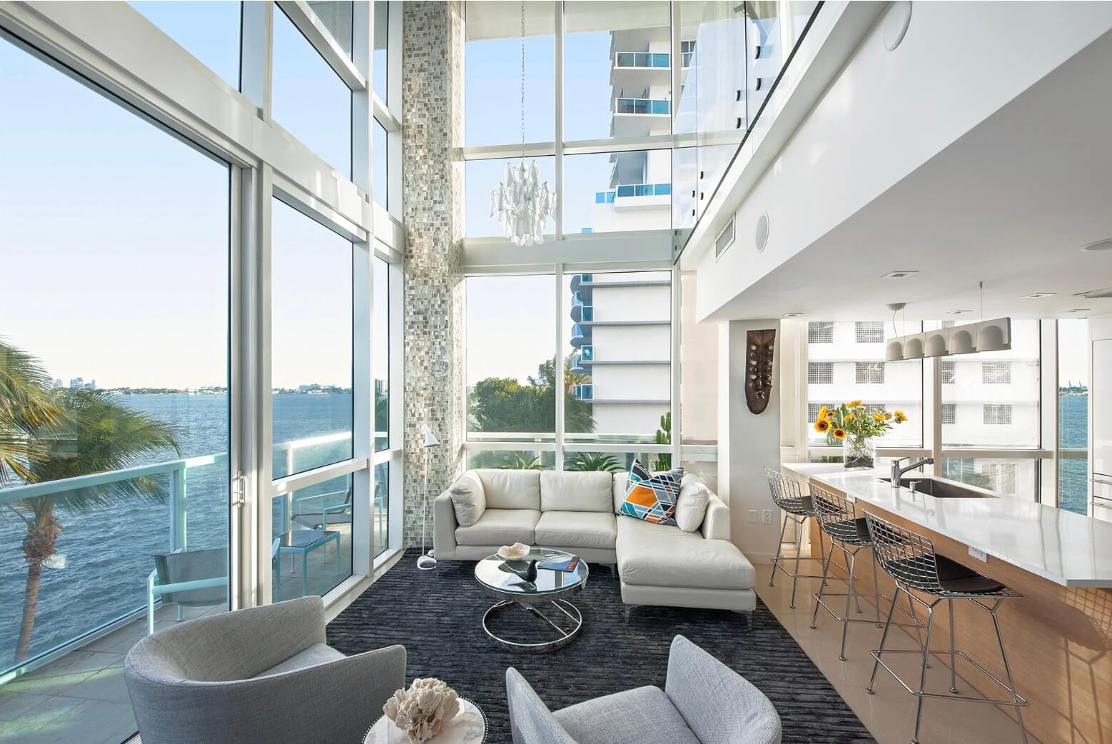 Double-height waterfront living space with white sectional, glass walls, modern kitchen island, and bay views