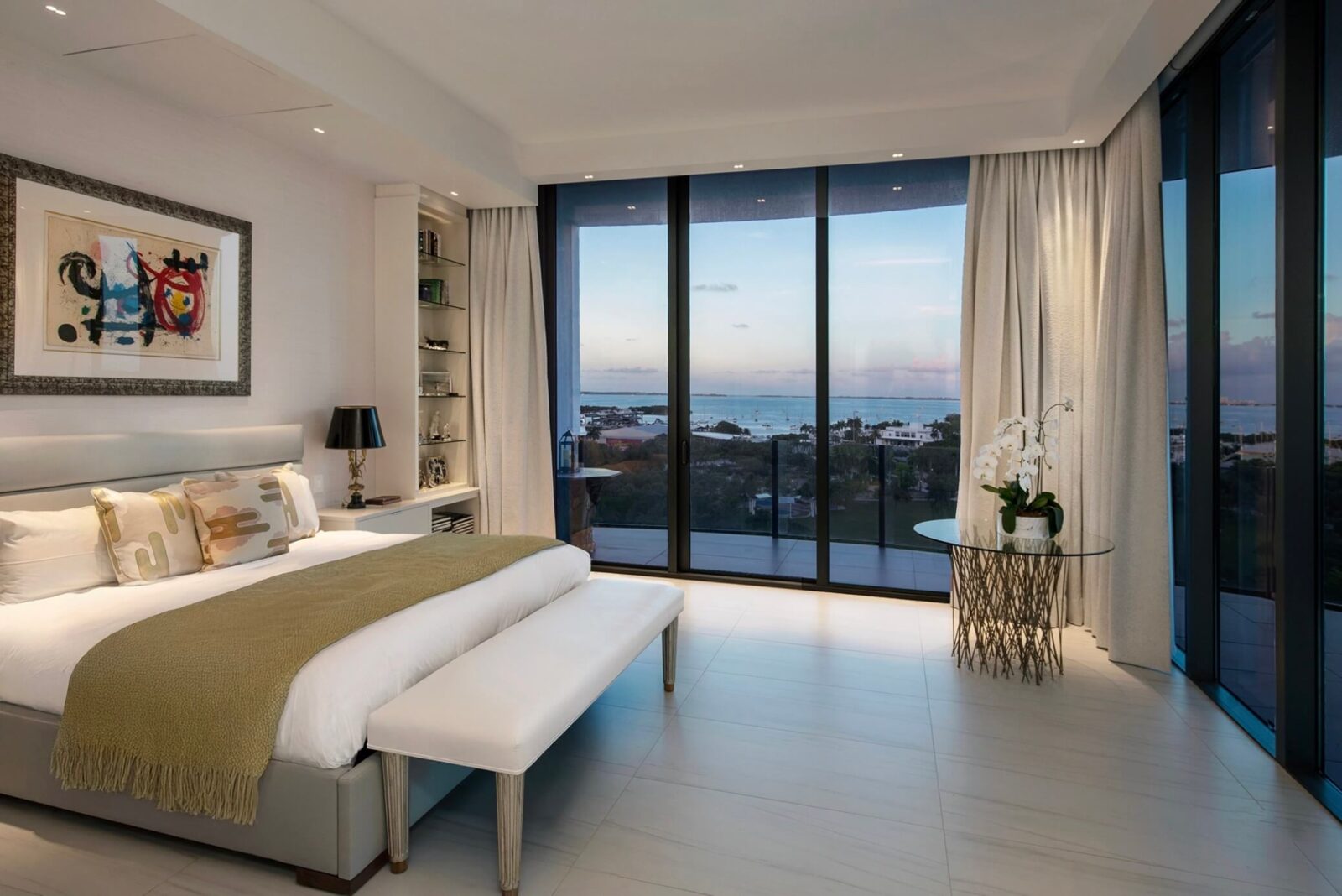 Minimal contemporary bedroom with upholstered bed, soft neutral textiles, and floor-to-ceiling windows overlooking the water