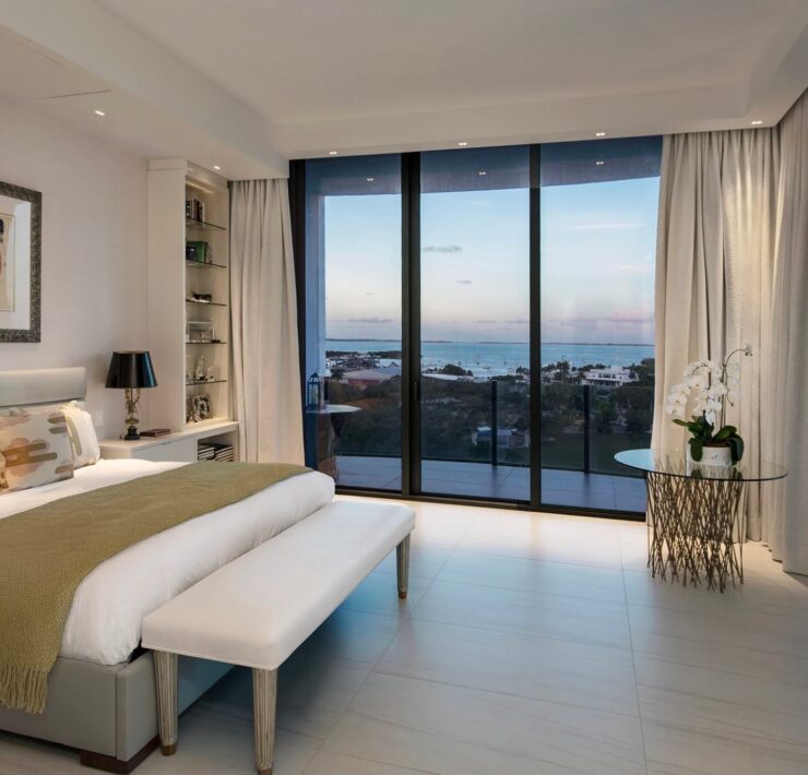 Minimal contemporary bedroom with upholstered bed, soft neutral textiles, and floor-to-ceiling windows overlooking the water