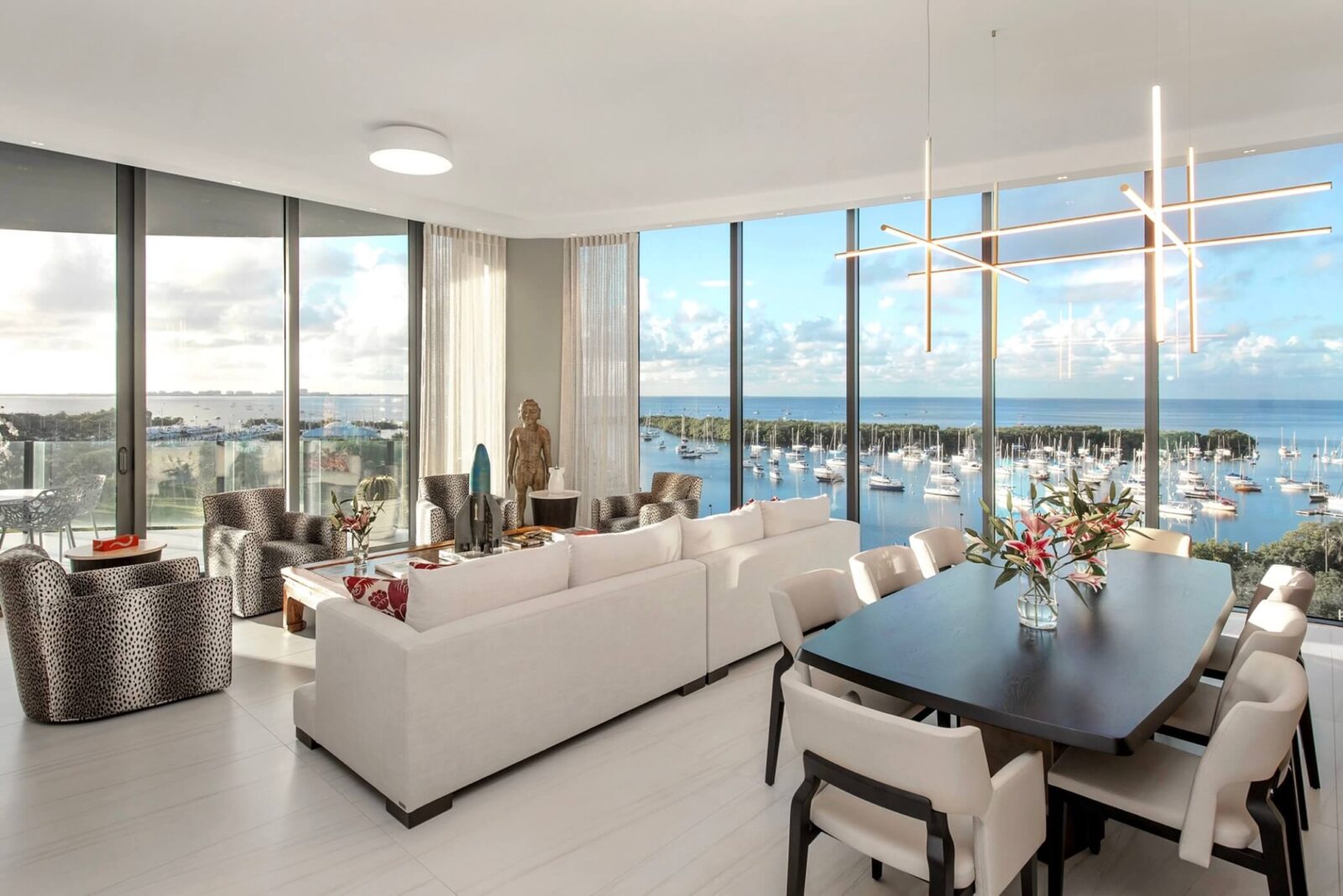 Bright modern living and dining room with marina views, white seating, sculptural chandelier, and black dining table