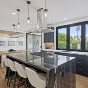 Modern kitchen with dark cabinetry, dramatic stone waterfall island, pendant lights, and large black-framed windows