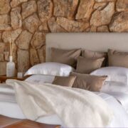 Trousseau Cairo Collection bedding styled in a neutral bedroom with white linens, stone wall, and warm wood accents