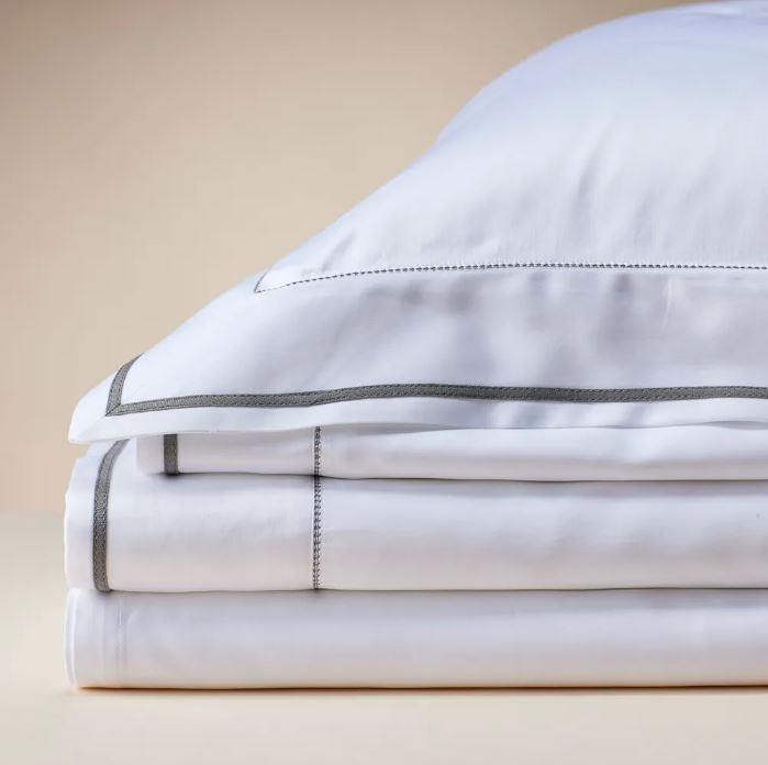 Trousseau Garda Collection bedding styled with white sheets, neutral pillows, and a soft beige throw